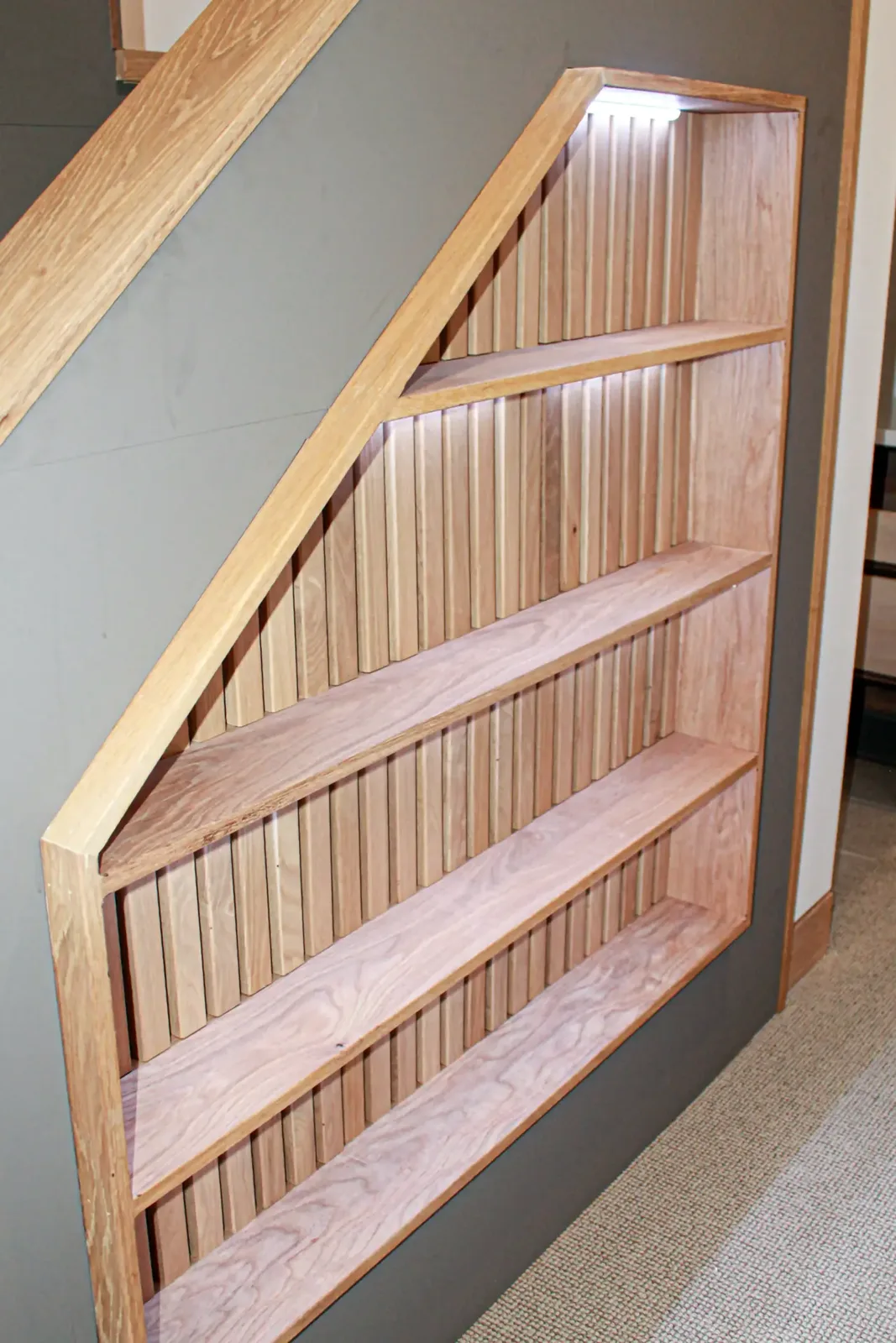 Angled built-in shelving unit set into a wall under a stair, with a fluted back panel and a strip light at the top
