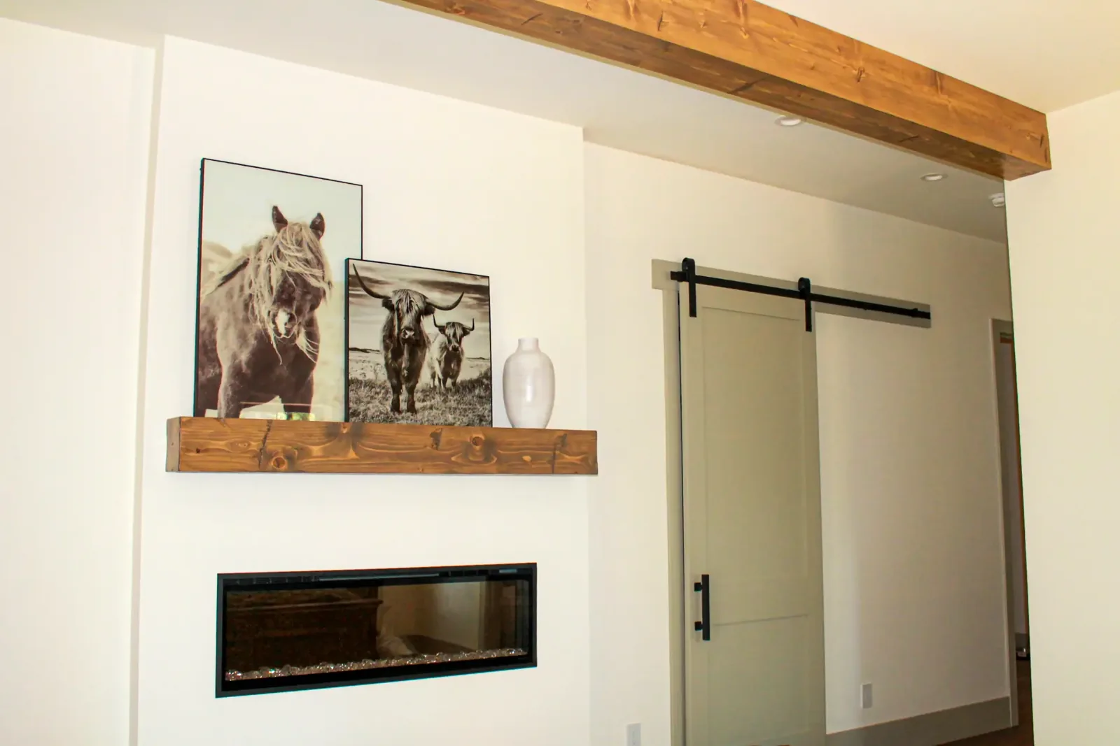 Wood floating mantel shelf above a linear fireplace, with a ceiling beam and sliding barn door nearby