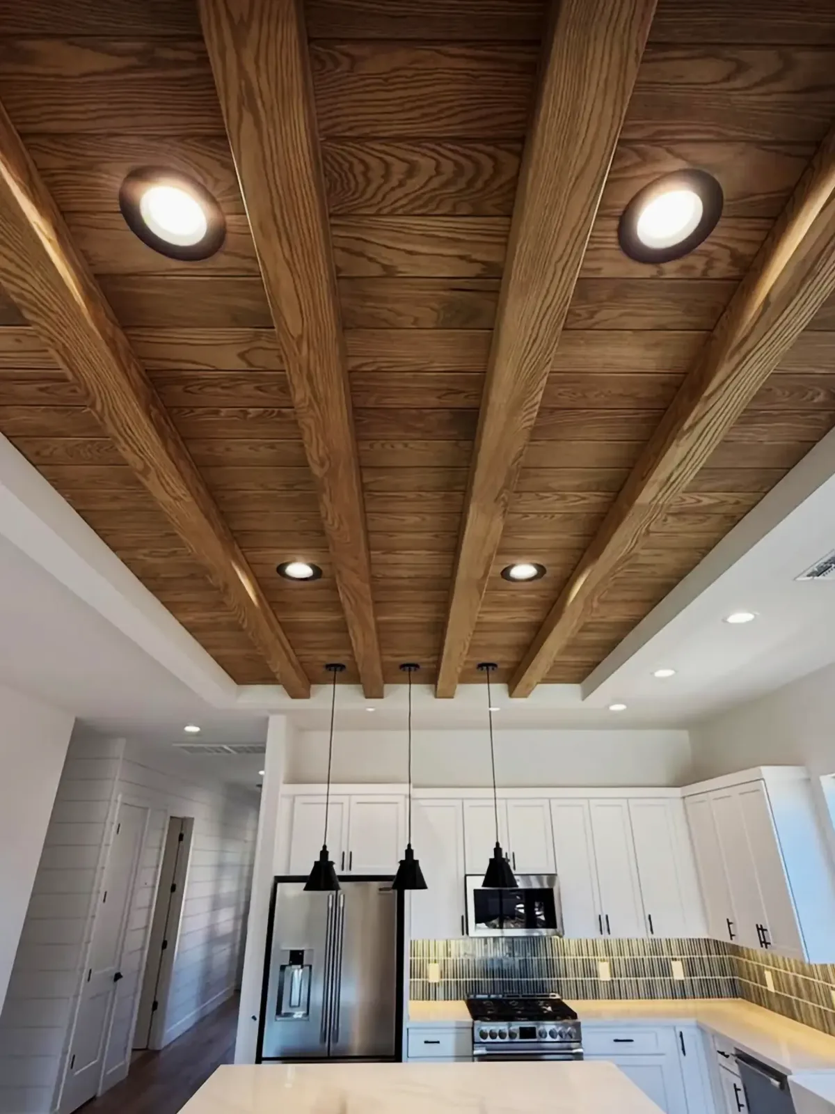 Recessed ceiling clad in wood planks with wood beams and recessed lights above a white kitchen