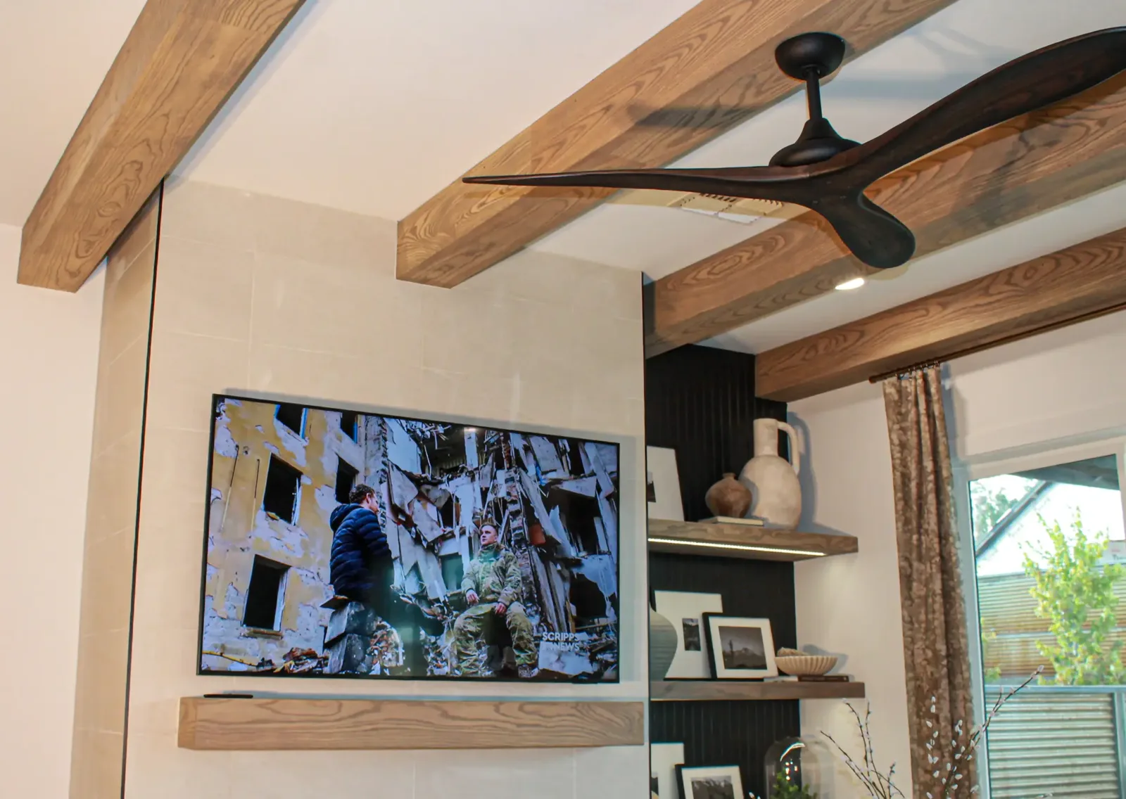 Living area with wood ceiling beams, a black ceiling fan, and a wood shelf mounted below a wall-mounted TV