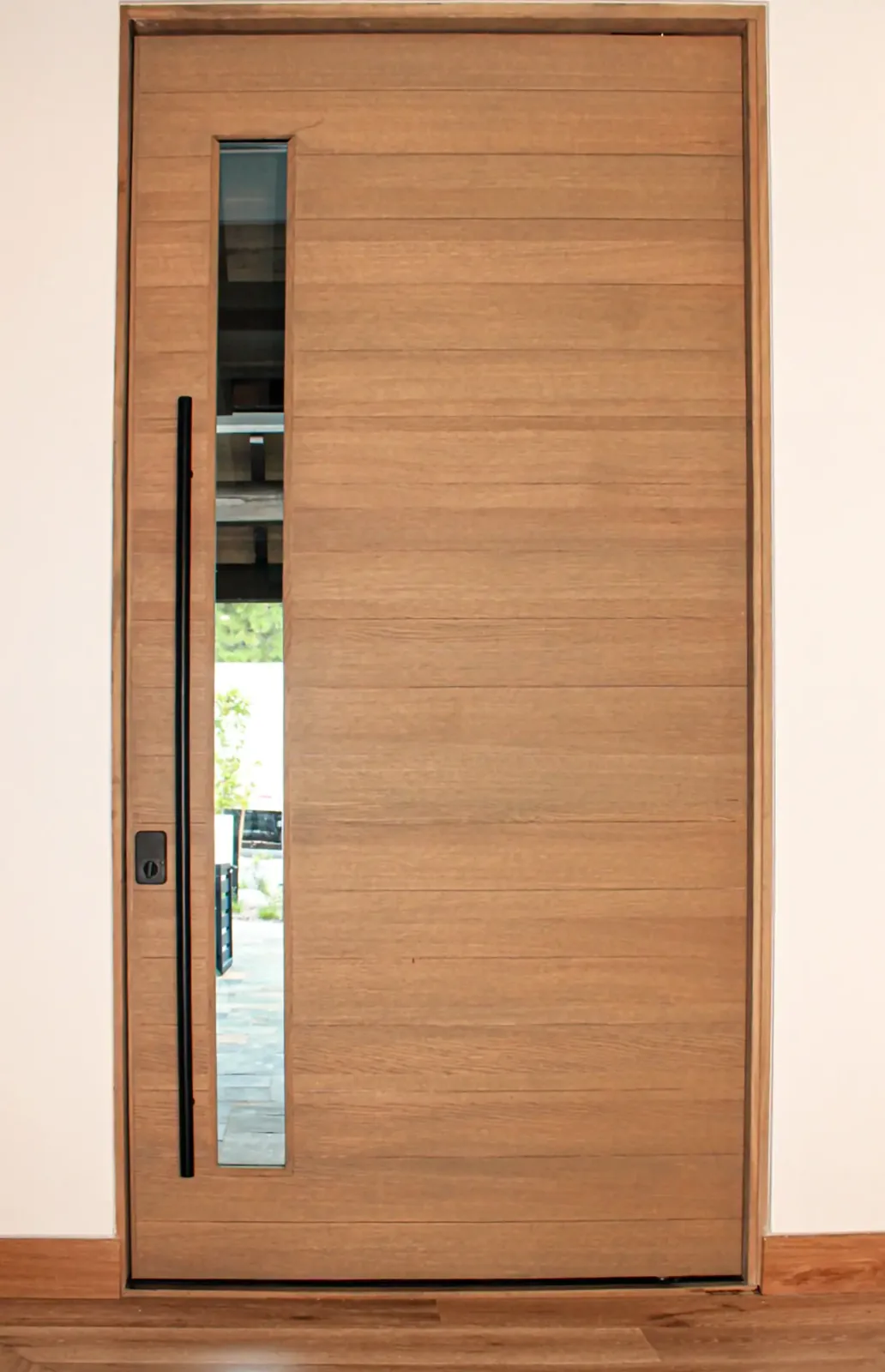 Flush wood entry door with a narrow vertical glass lite, long black pull, and matching wood jamb