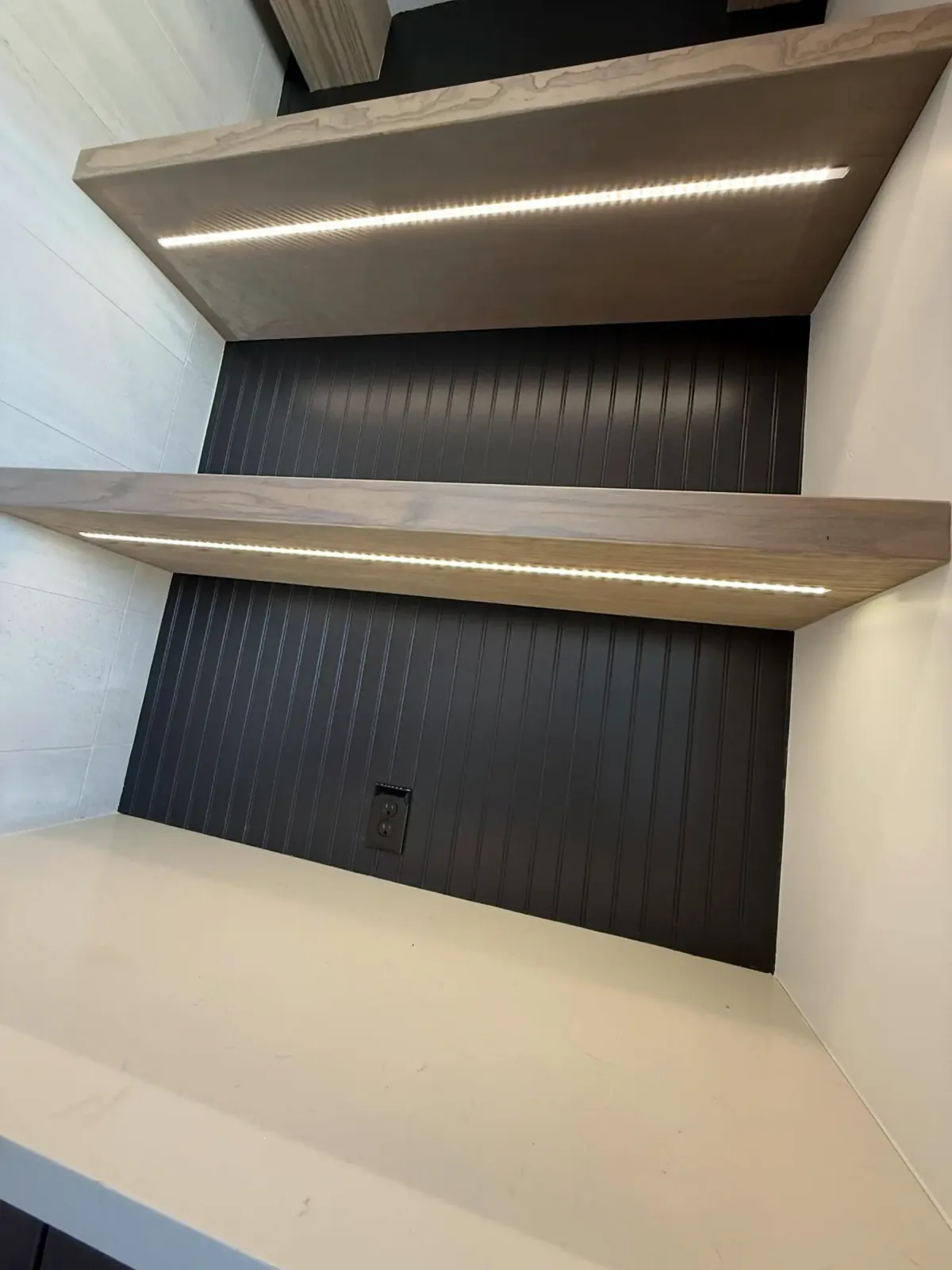 Two wood floating shelves with LED strips on their undersides above a black beadboard back panel
