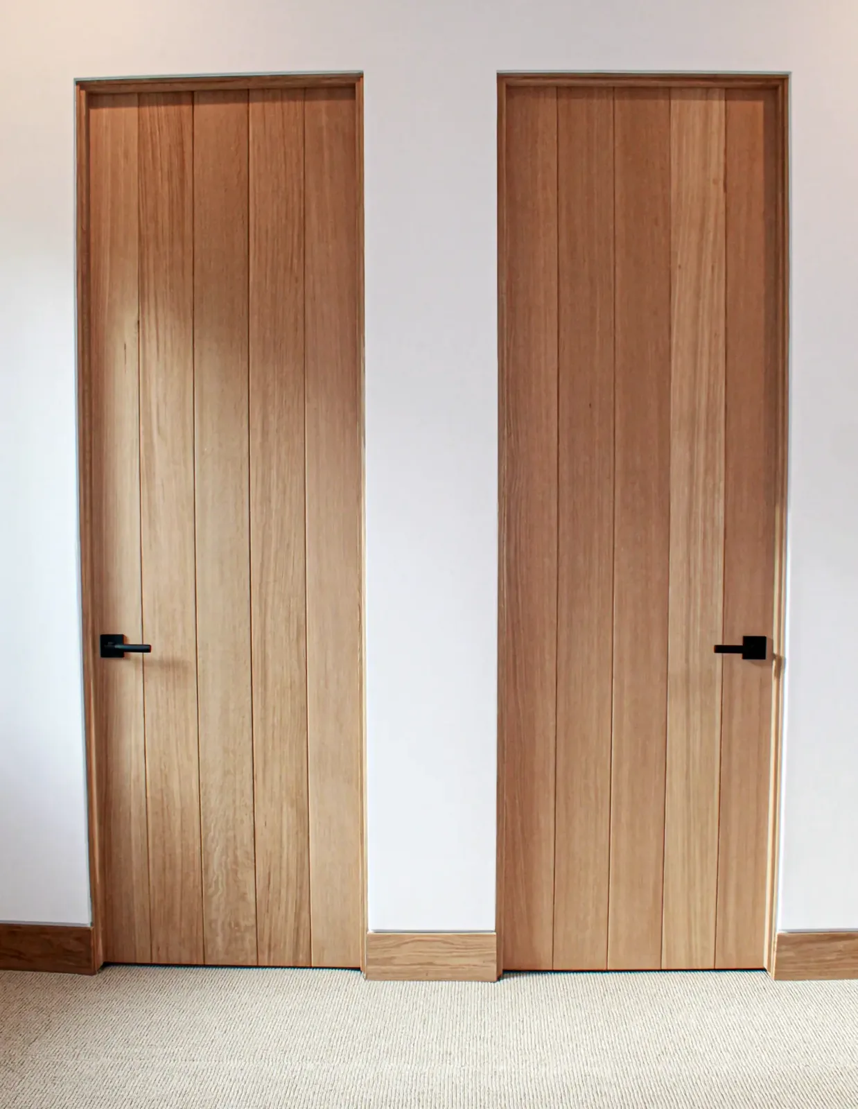 Two flush wood doors with vertical plank faces and black lever handles, set in matching wood jambs