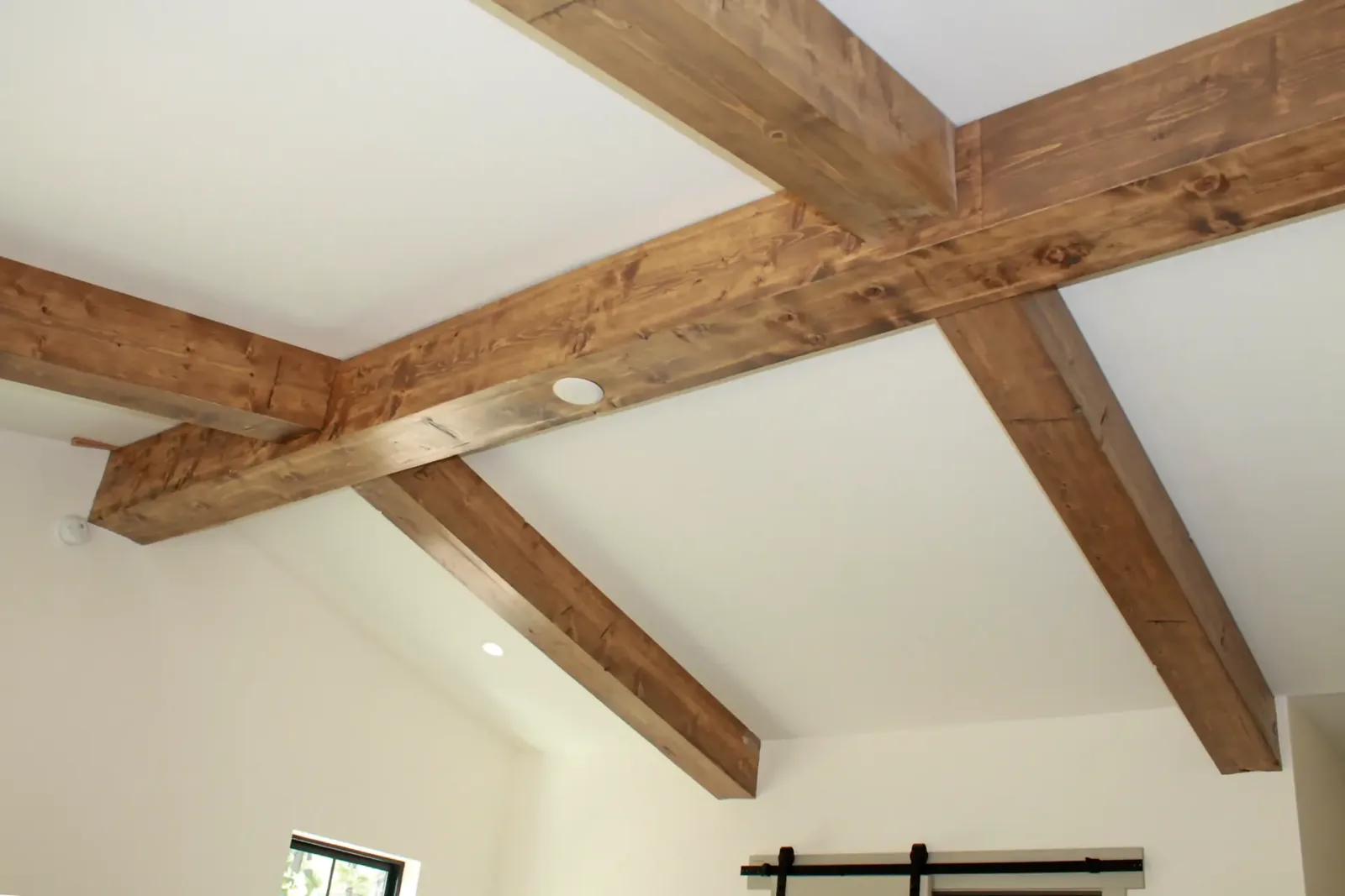 Stained ceiling beams meeting at a cross joint above a white wall and window