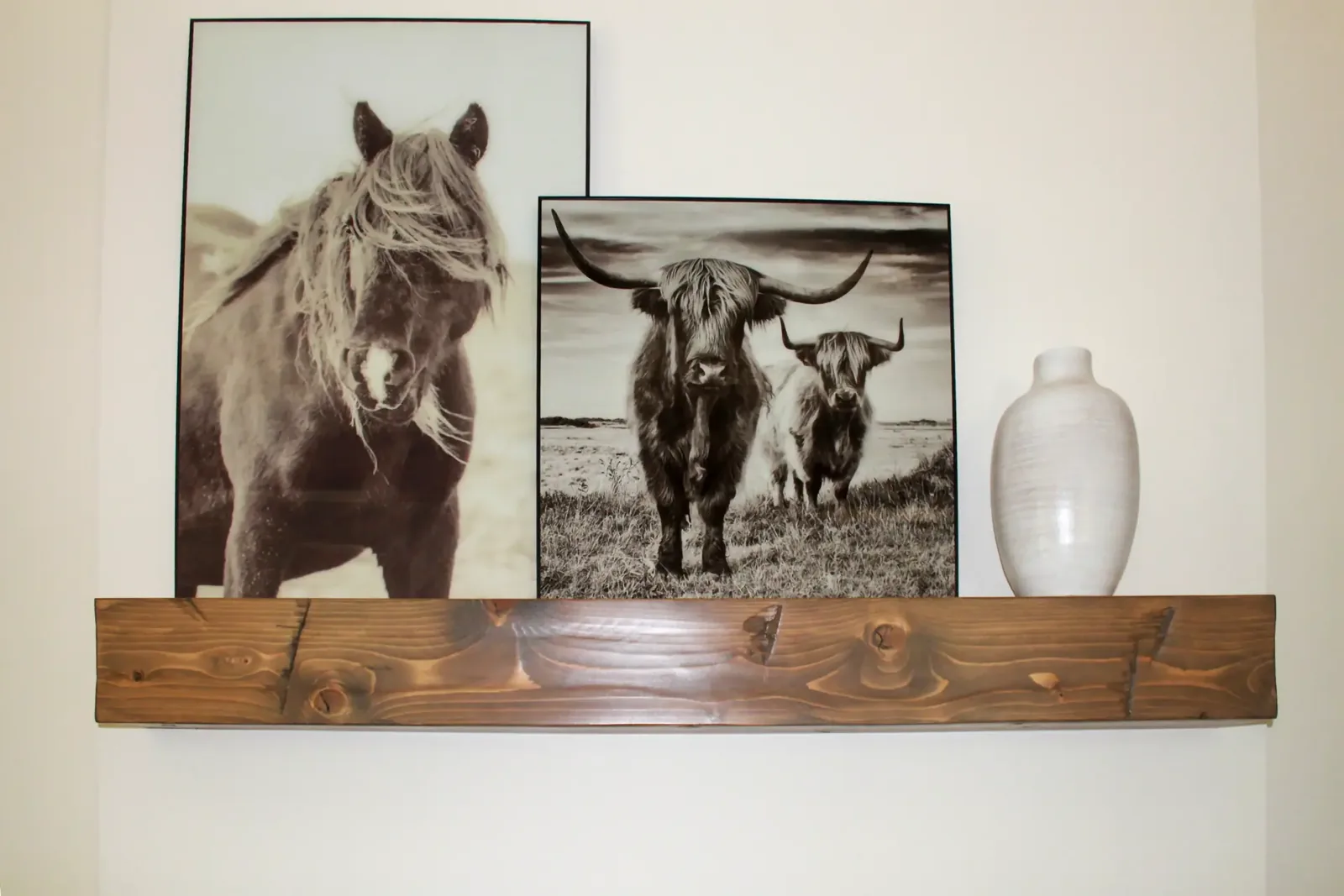 Close view of a wood floating mantel shelf holding two framed prints and a white vase