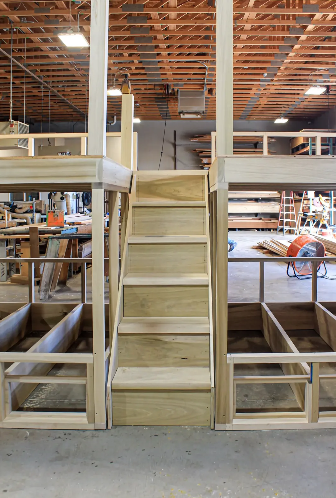 Shop view of a stair assembly flanked by unfinished cabinet frames, with lumber racks in the background